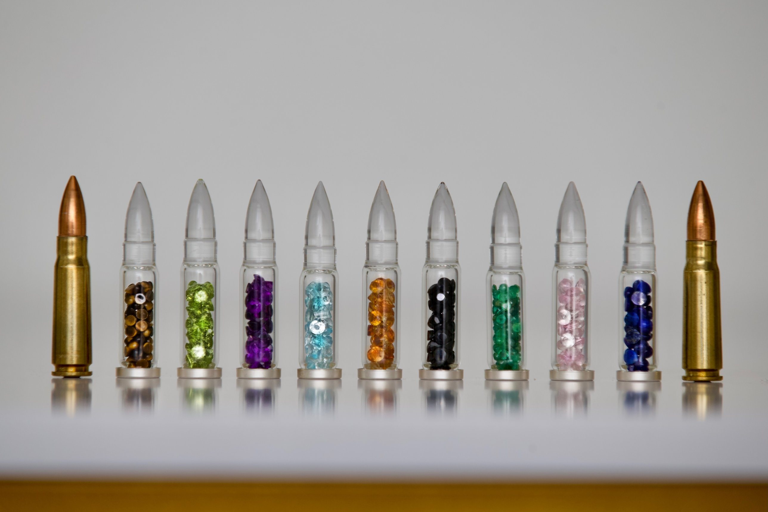 A row of hand-blown glass bullets filled with coloured gemstones, between two brass AK-47 rounds